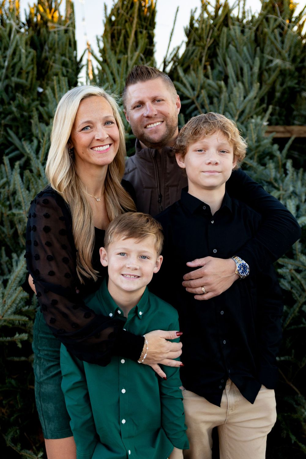 Family poses together in front of evergreen Christmas trees during holiday photo session.