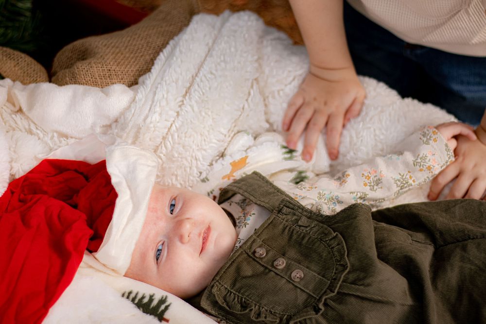 A baby wearing military-style clothing lies on a white blanket with red holiday decor.