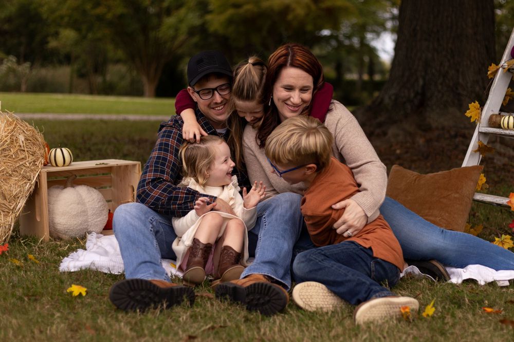 Family lounging together on a blanket outdoors with hay bales and fall decor in a park setting.