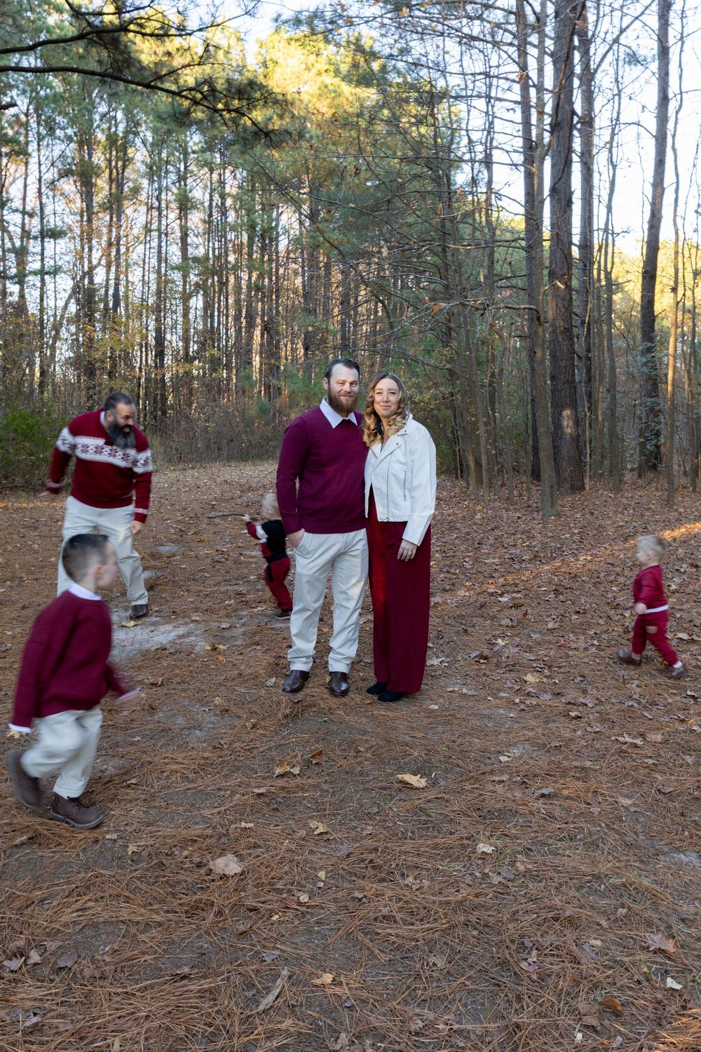 A family in coordinating burgundy and white outfits poses together on pine needle covered ground in a wooded forest setting.