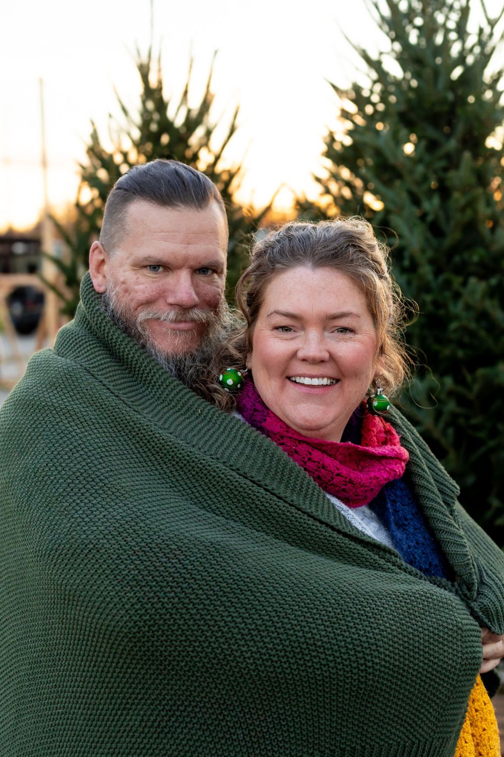 Couple wrapped in a cozy green blanket smiling together among evergreen trees at dusk.