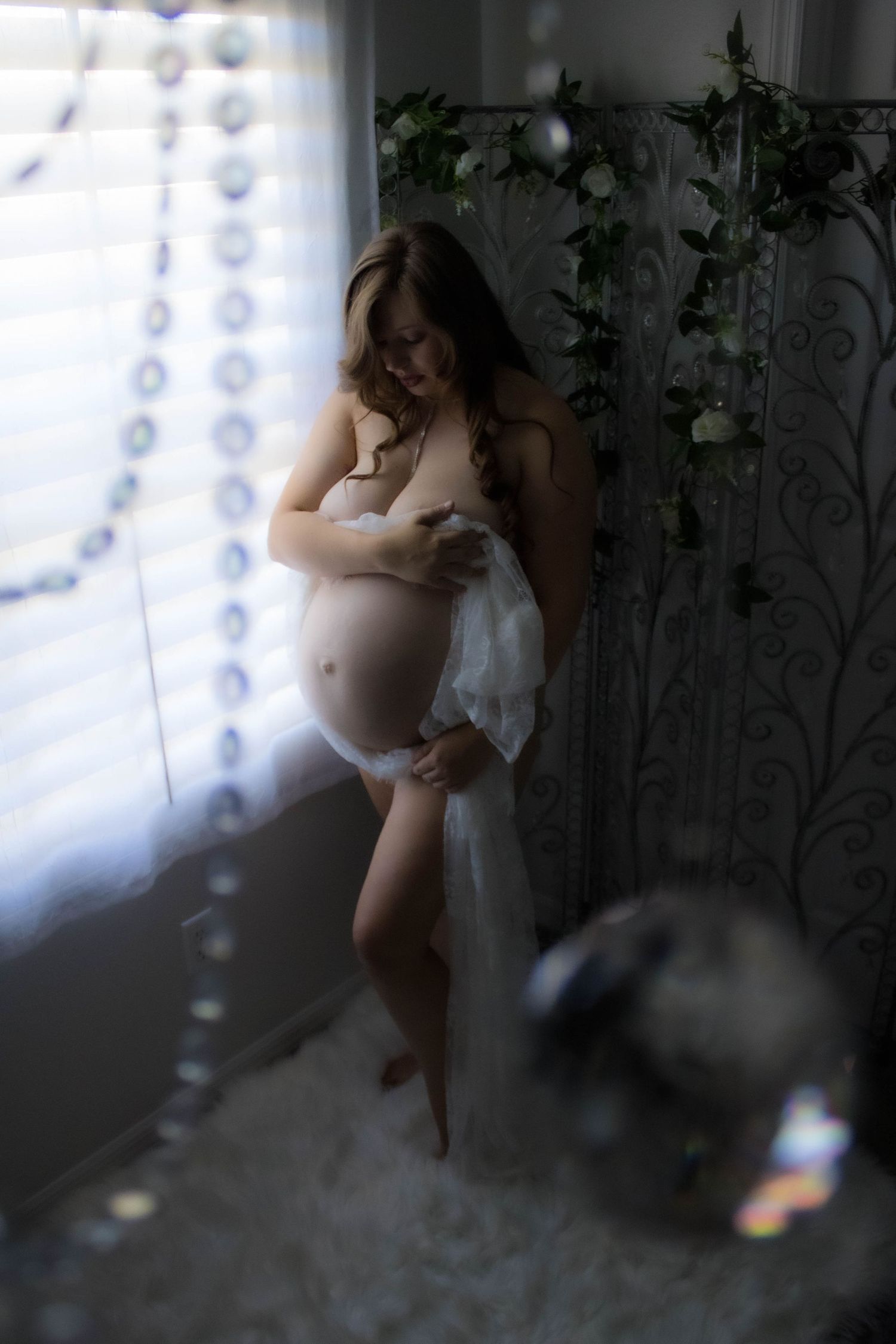 A dreamy maternity photo by a window with sheer curtains and indoor plants in soft natural lighting.