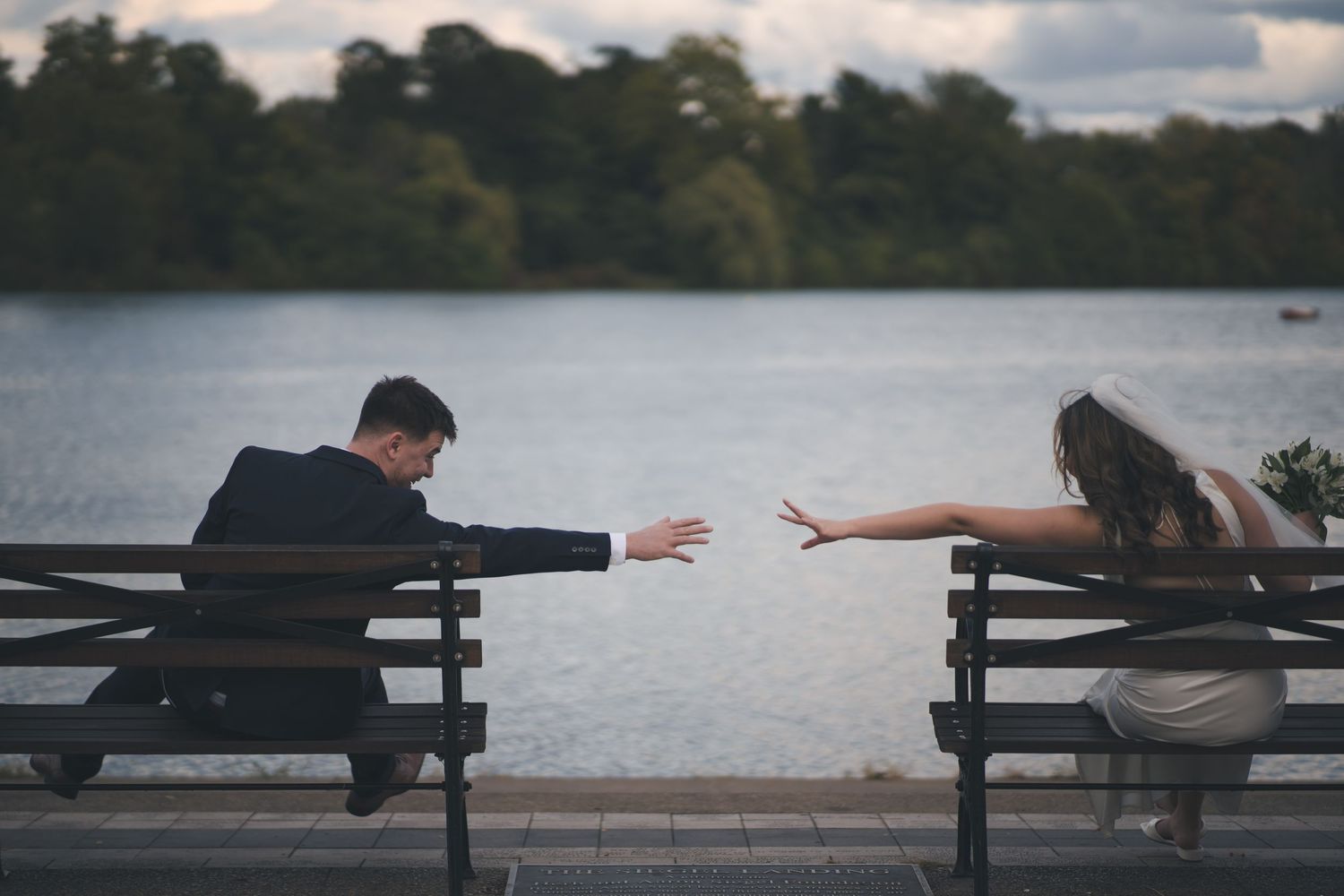Two people reach toward each other while sitting on separate benches by a lake.