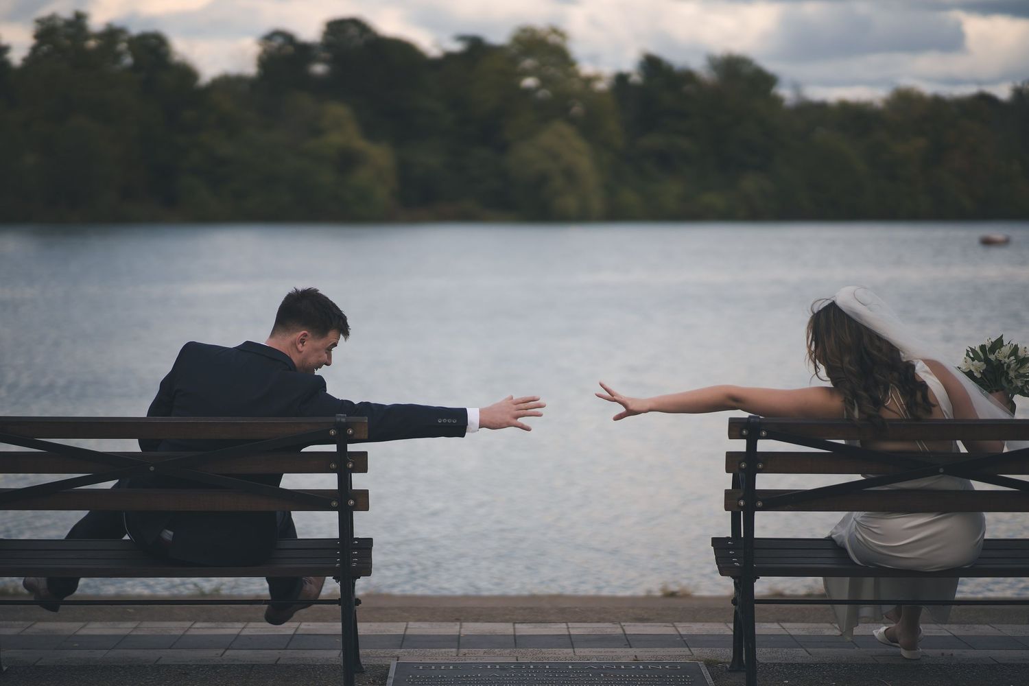 Buffalo NY wedding bride and groom reach toward each other while sitting on separate benches by a lake.