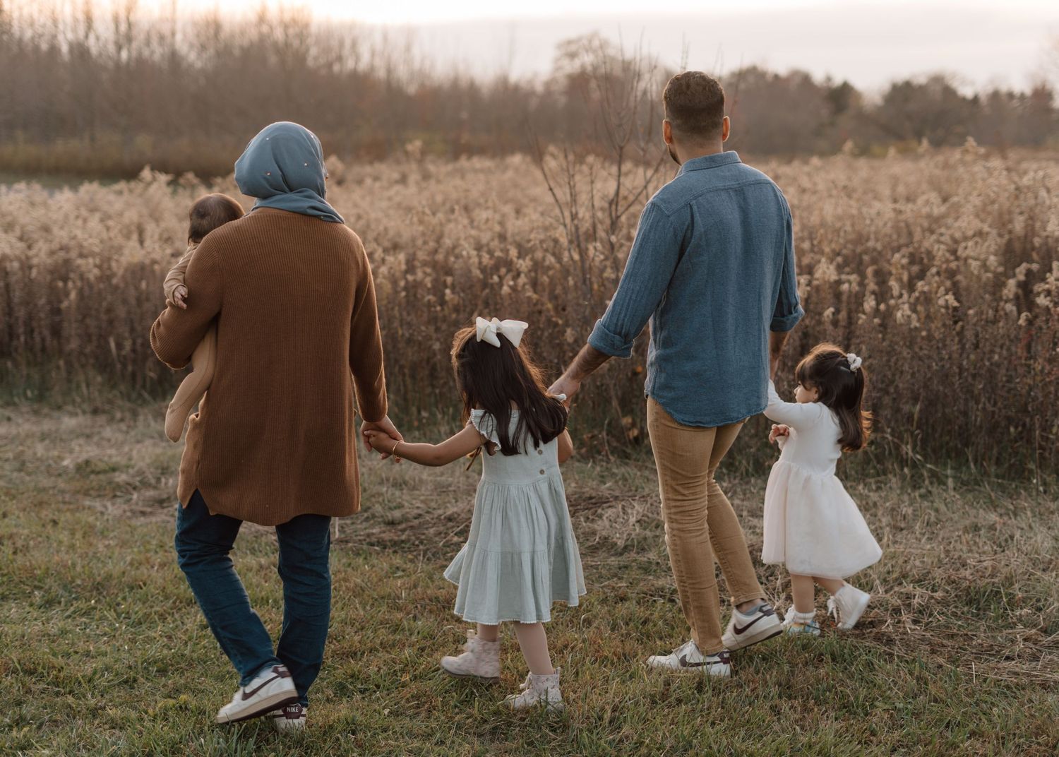 Family walking together holding hands with two children in white dresses through autumn field.