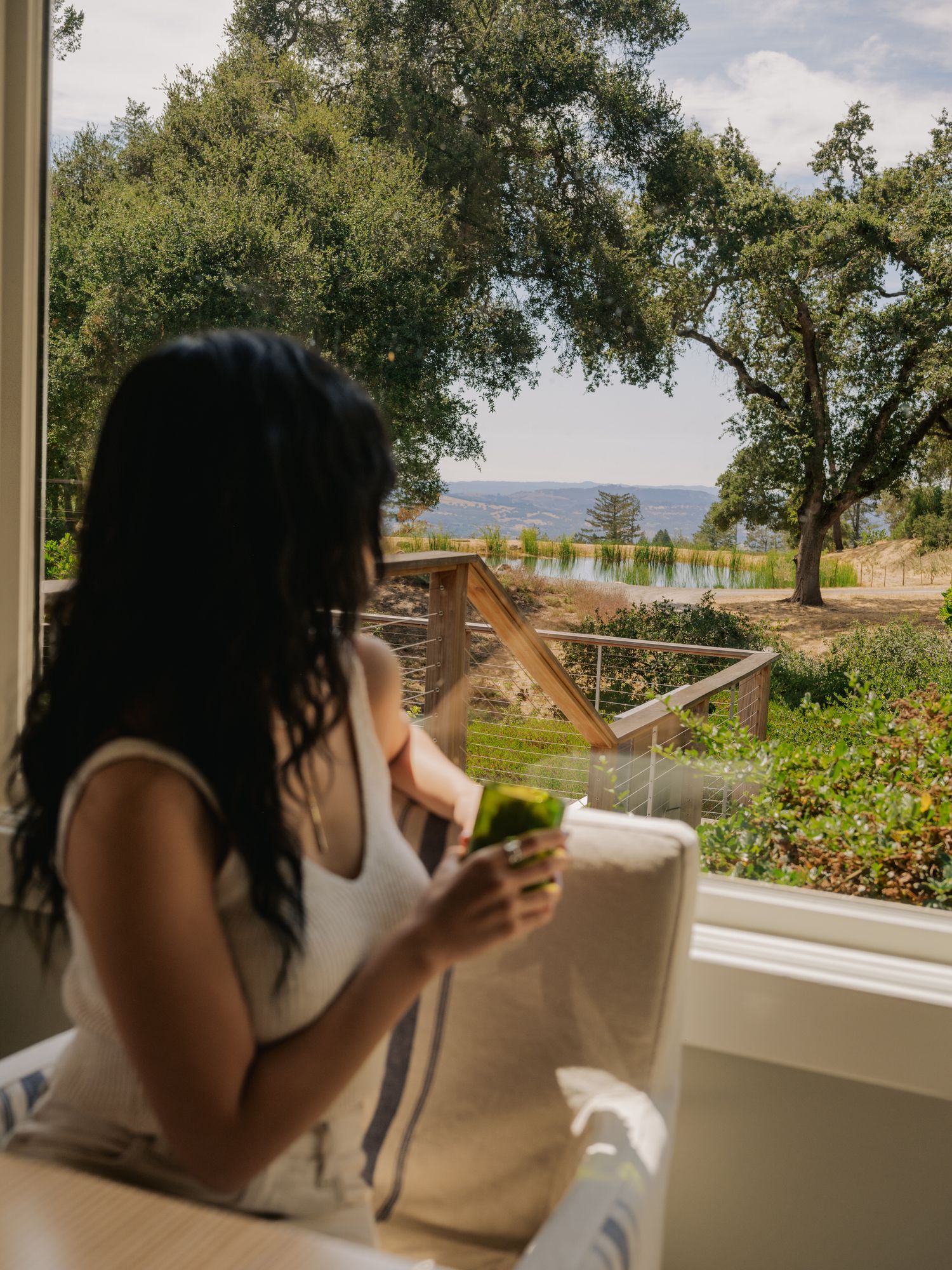 Person enjoying a beverage on a porch overlooking a scenic lake with trees in golden afternoon light.