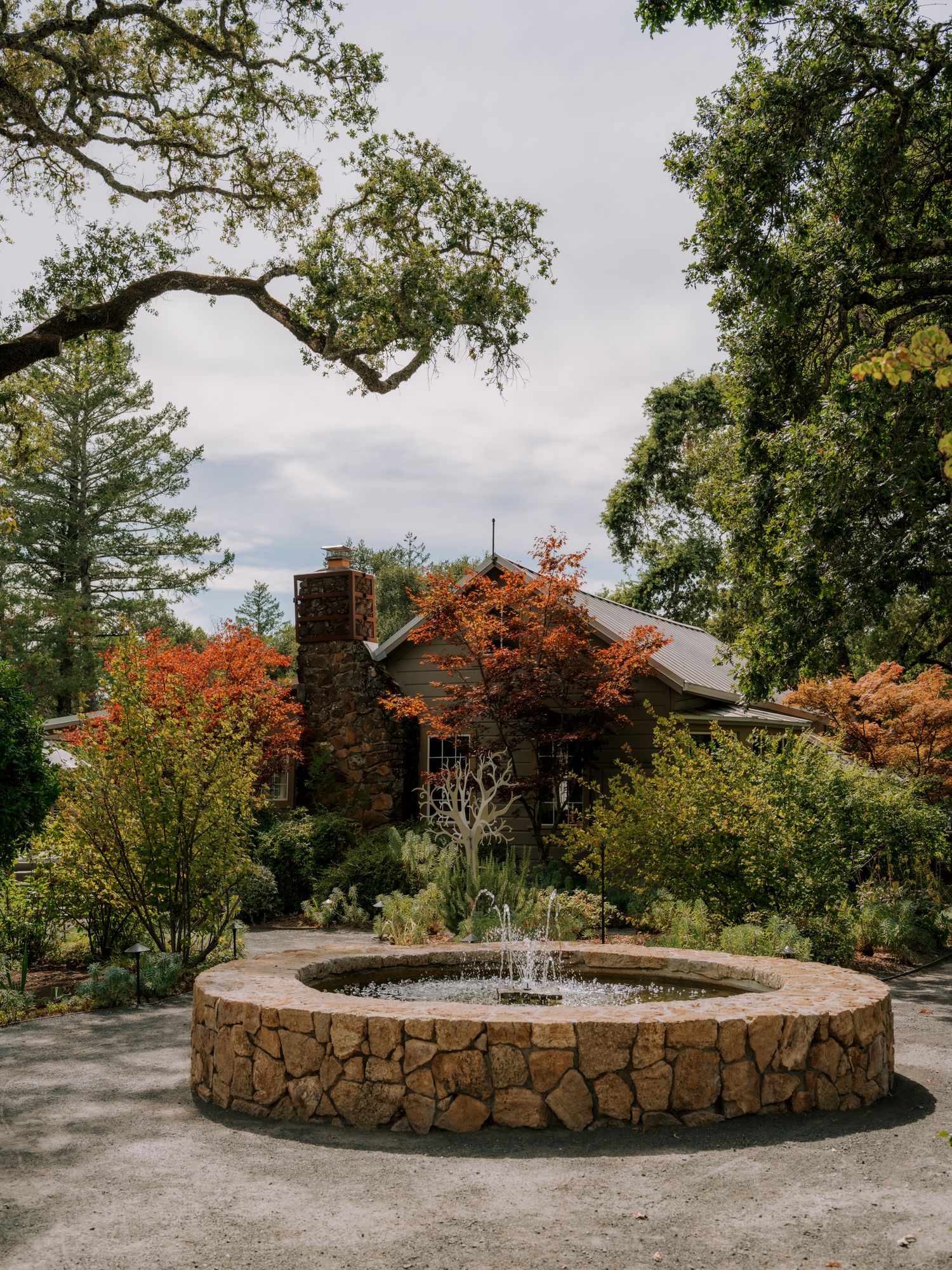 A stone circular fountain with water feature sits in front of a rustic house surrounded by autumn trees and foliage.