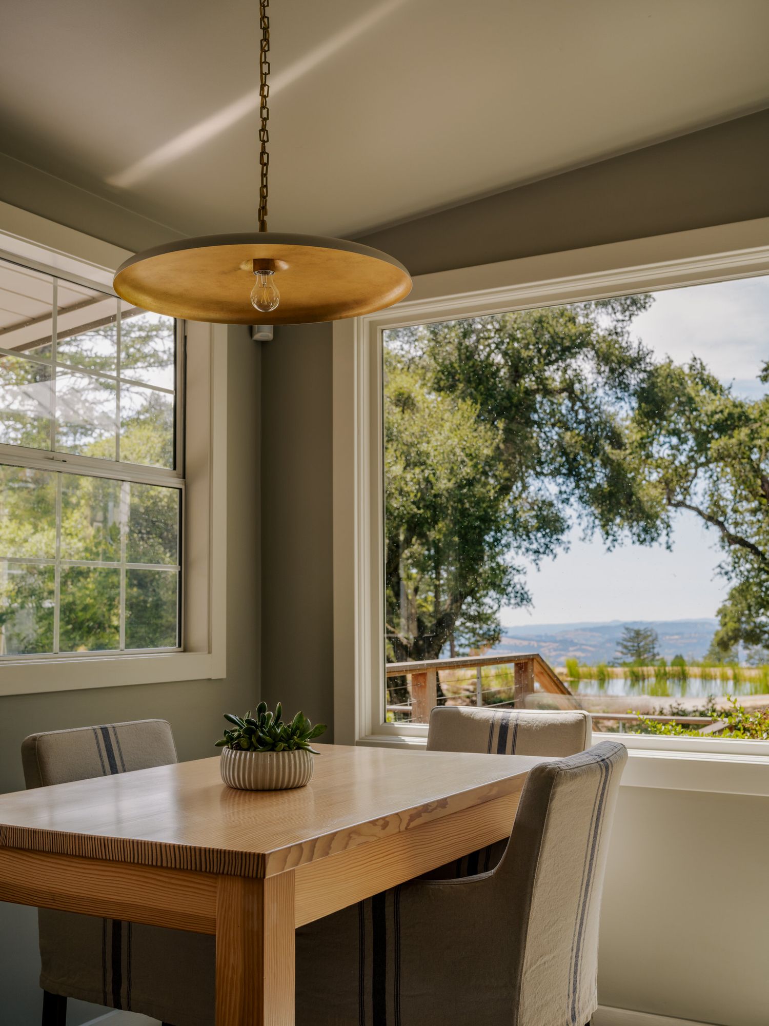Modern dining area with wood table and gray chairs features a UFO pendant light and large windows overlooking a waterfront view.