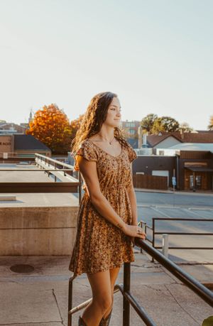 Standing on a rooftop terrace in a brown floral dress against an autumn cityscape background.