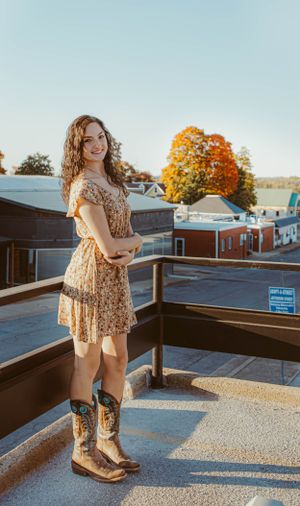 Wearing cowboy boots and a floral dress on a rooftop during golden hour with fall foliage visible.