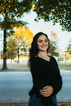 Standing in front of fall trees wearing black sweater and jeans on a sunny afternoon.