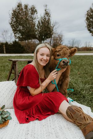 A red dress-clad woman sits on a blanket with a baby cow wearing a turquoise halter.