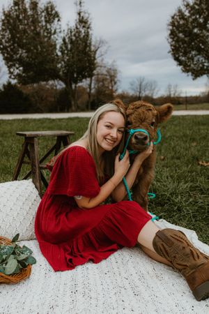 Woman in red dress cuddling with young calf on white blanket in grassy field.