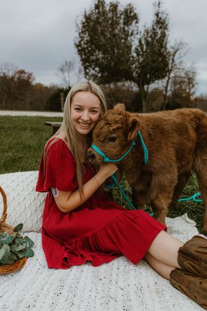 Woman in red dress petting brown calf with turquoise halter on white blanket.