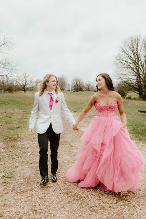 A formal couple in a white suit and pink ball gown walk together on a dirt path.