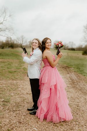 Two people pose back-to-back with a white suit and pink ball gown in a rustic outdoor setting.