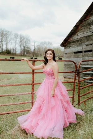 A person in a flowing pink ball gown poses near a rustic barn and metal gate.