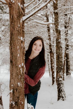Person peeking from behind tree trunk in snowy winter forest.