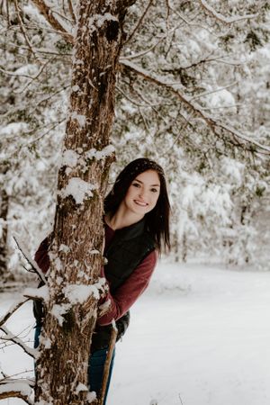 Person peeking playfully from behind a snow-covered tree trunk in a winter forest setting.