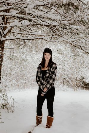 Person wearing plaid flannel and beanie poses in snowy winter forest landscape.