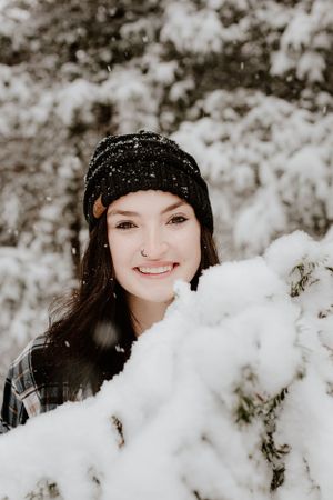 Winter portrait with person wearing knit beanie among snow-covered evergreen branches.