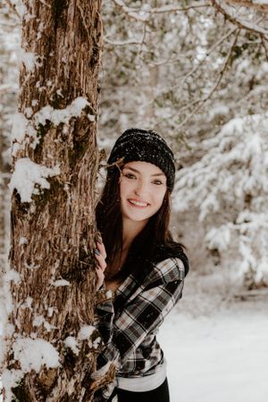 Person in plaid shirt and beanie poses by snow-covered tree trunk in winter forest.