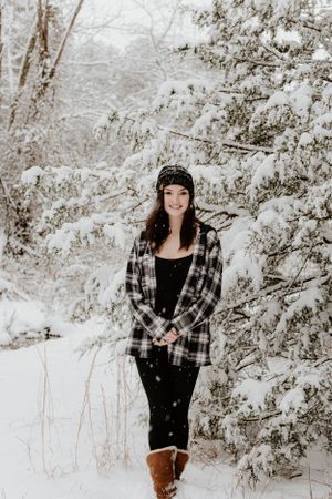 Full-length portrait of person in plaid and boots standing in snowy winter forest.
