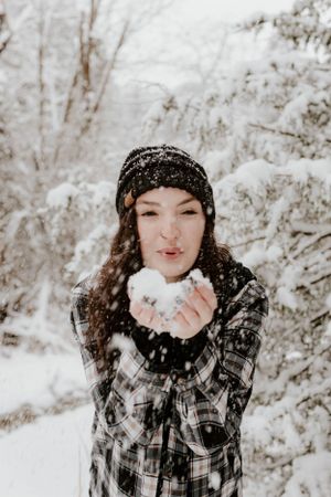 Person in plaid shirt blowing snow from hands in winter forest setting.