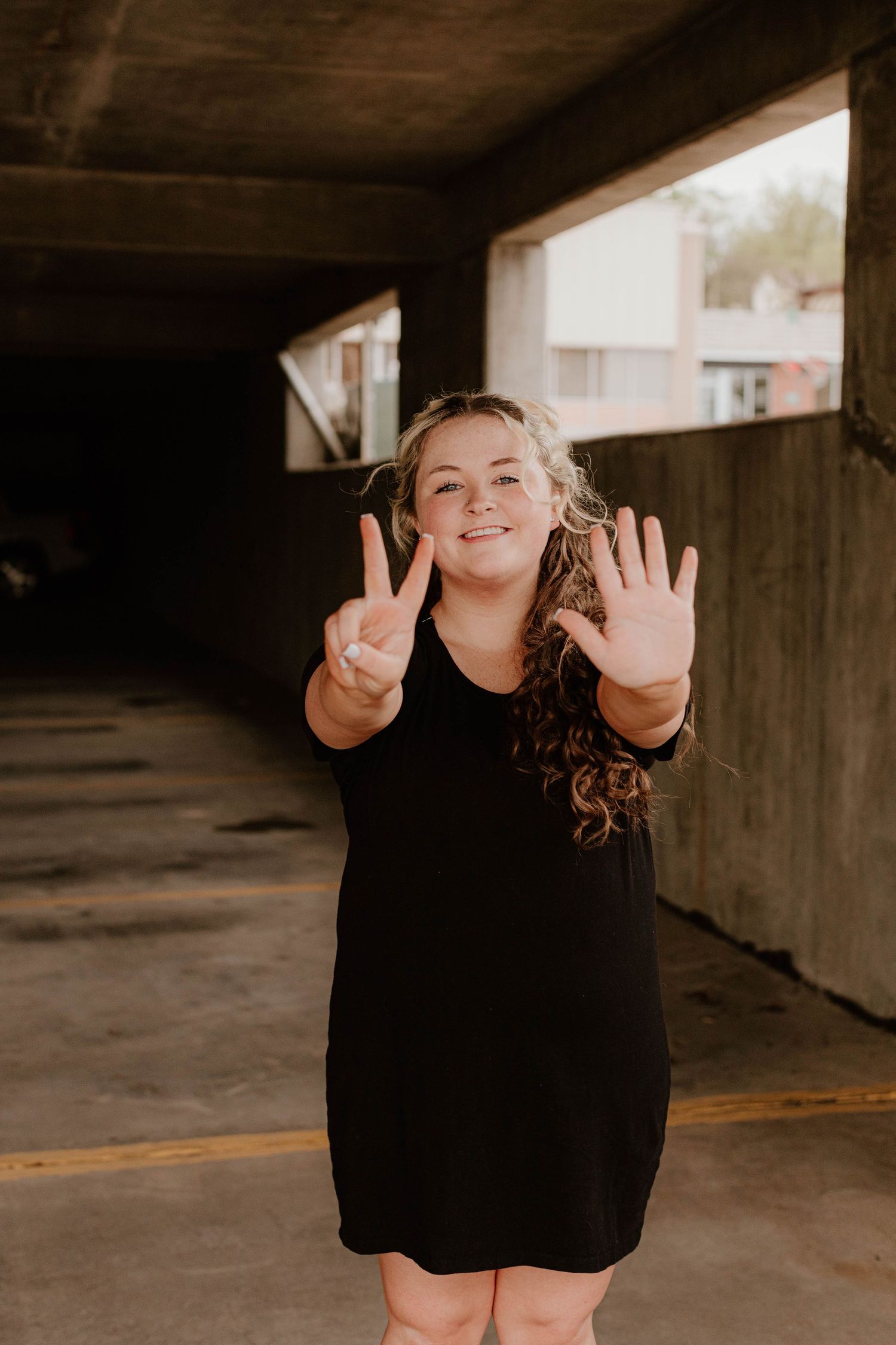 A person in a black dress making peace sign and waving at the camera in a parking garage.