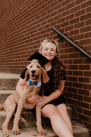 A happy person in black dress posing with their golden retriever wearing a graduation cap.