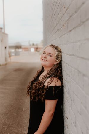 A person in a black dress smiling while leaning against a white brick wall.