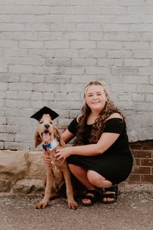 A person crouching next to a dog wearing a graduation cap against a white brick wall.
