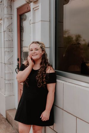 A person in an off-shoulder black dress posing against a white building exterior.