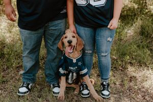Close-up of a happy dog in a sports jersey standing between two people's legs.