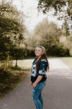 A person in sports jersey and jeans posing on a tree-lined path.