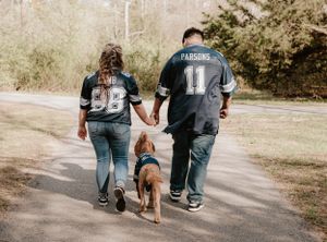 A couple in sports jerseys walking their dog on a nature trail holding hands.
