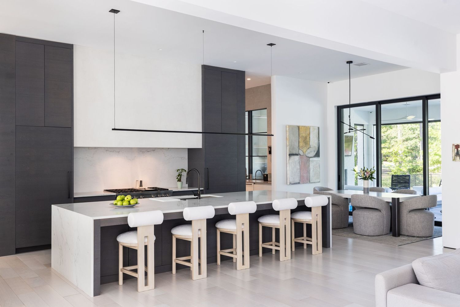 Modern kitchen with dark cabinetry, white island, bar stools, and open living area with large windows.