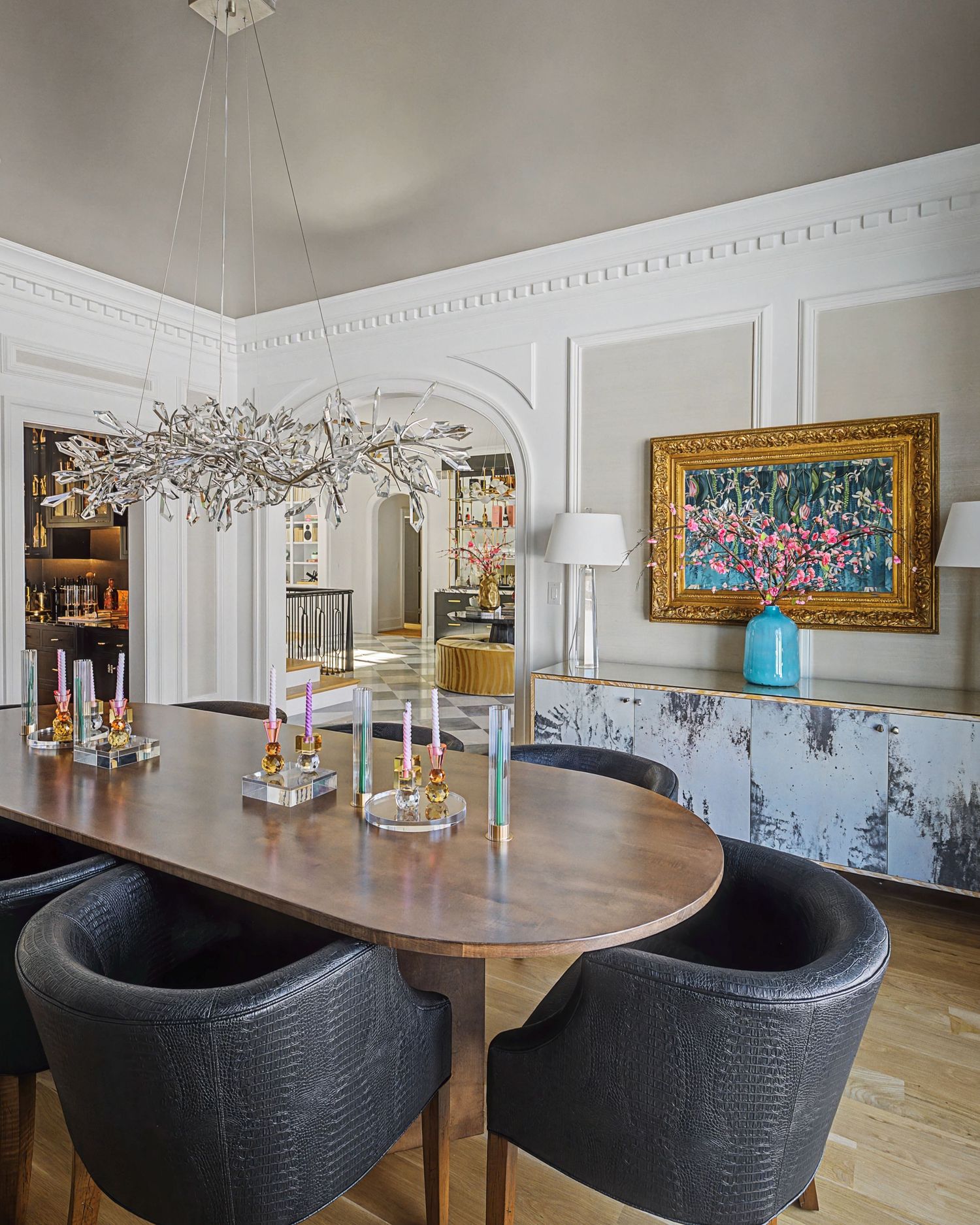 Elegant dining room with walnut table, dark leather chairs, crystal chandelier, ornate molding, and colorful floral artwork.