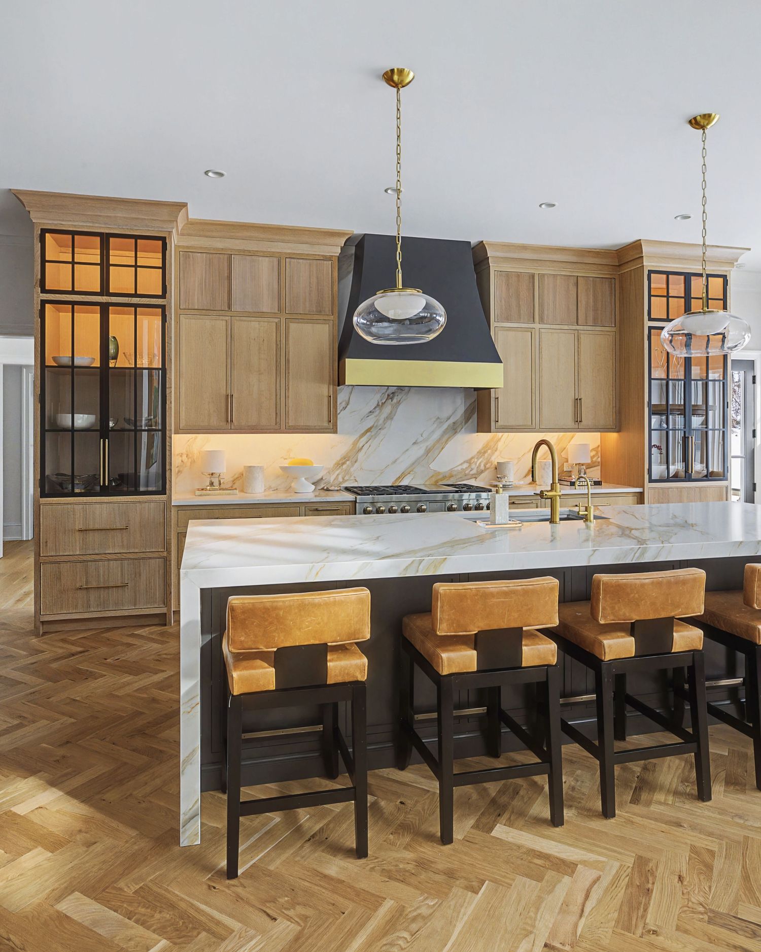 Elegant kitchen with oak cabinets, marble island, leather bar stools, black hood, brass accents, and herringbone hardwood floors.