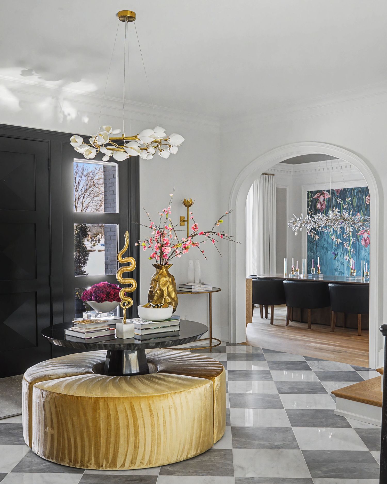 Elegant foyer with gold velvet ottoman, checkered marble floor, modern chandelier, and arched doorway revealing blue dining room.