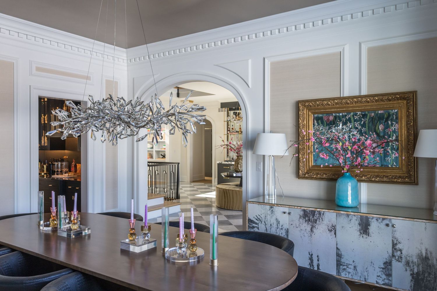 Elegant dining room with crystal branch chandelier, oval table, colorful accents, ornate moldings, and impressionist artwork on wall.