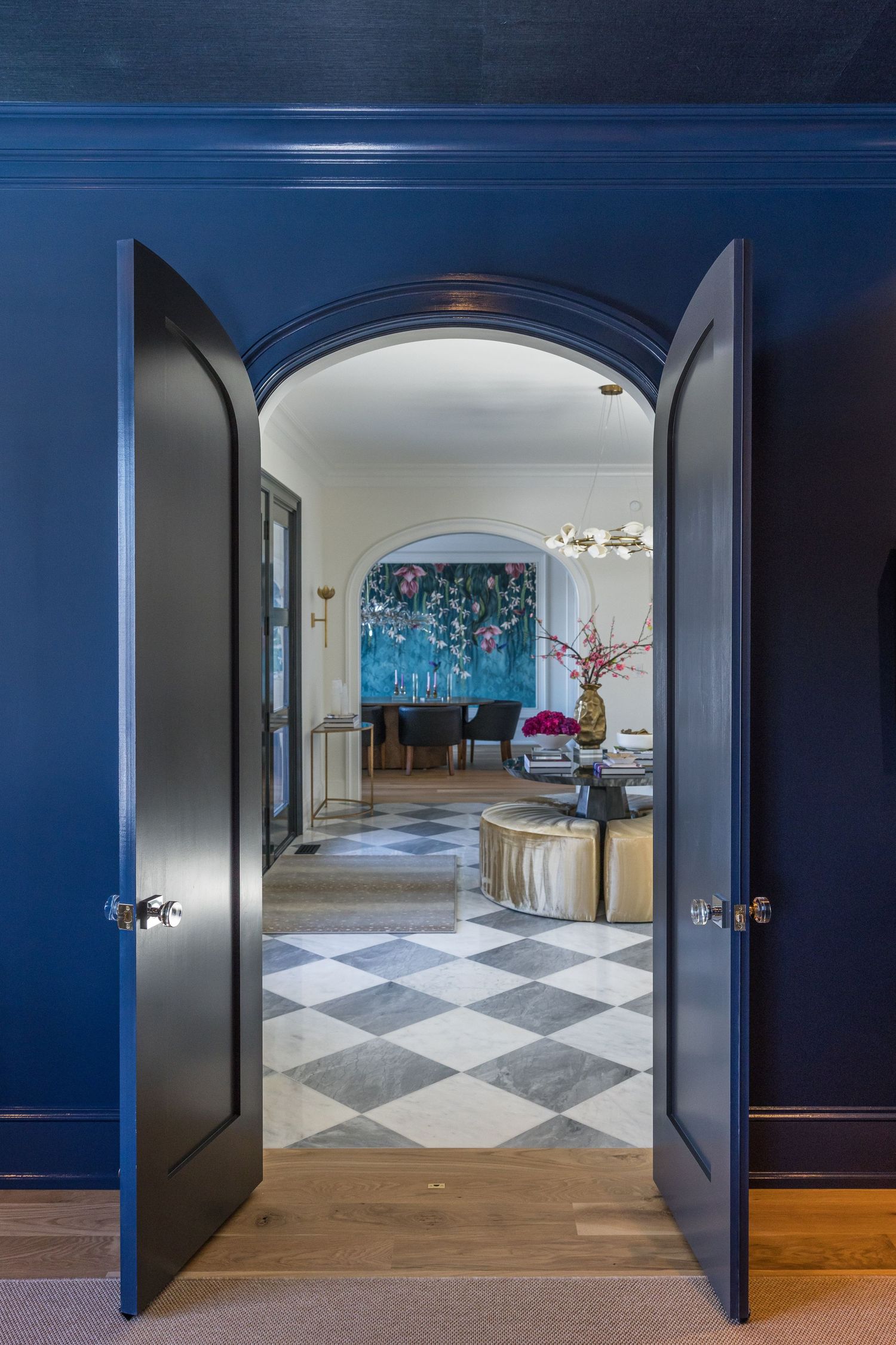 Navy blue double doors open to reveal a chic foyer with checkered marble floors and colorful floral wall art beyond.