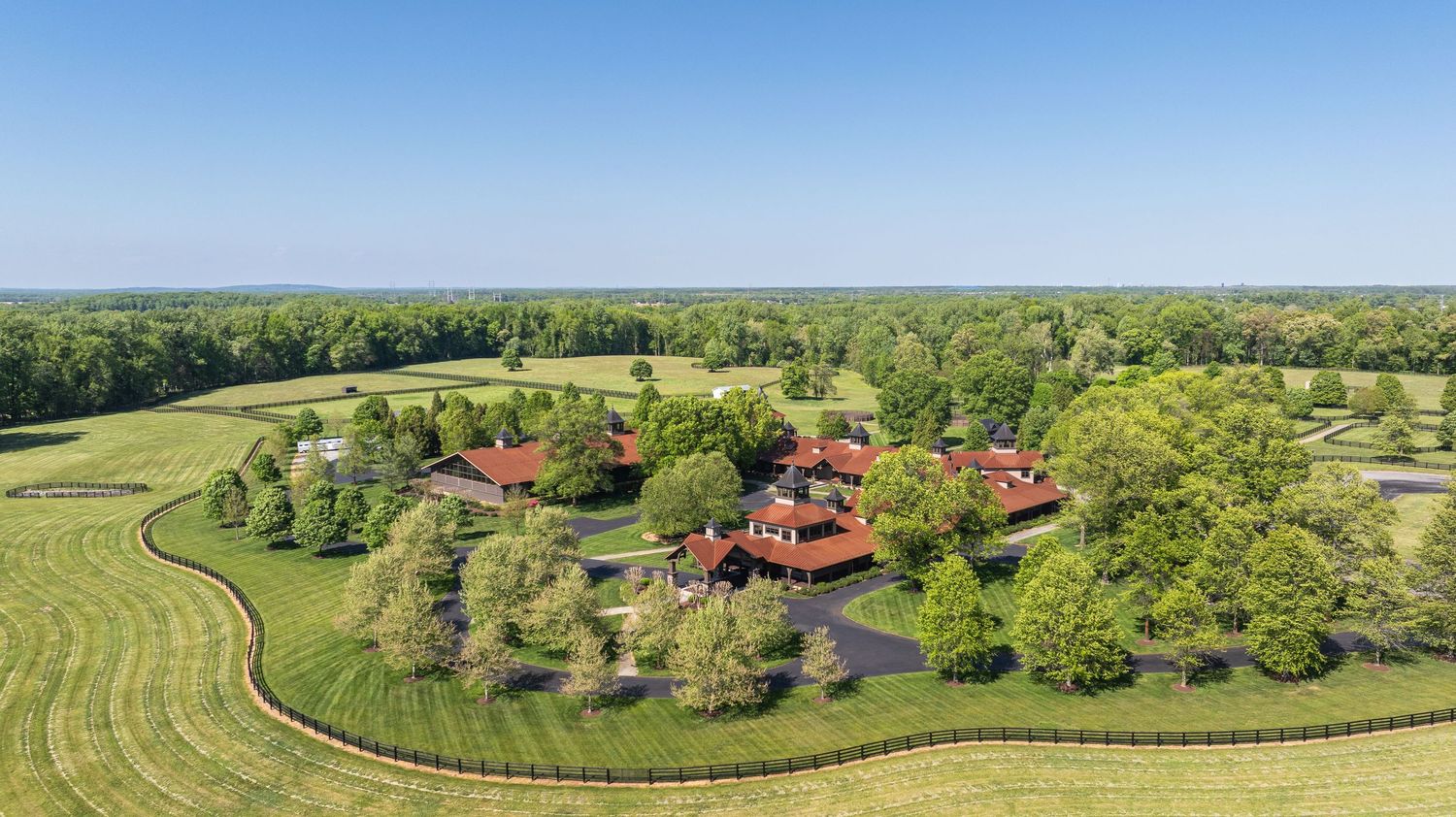 Aerial view of a sprawling red-roofed estate surrounded by lush green fields, manicured lawns, and mature trees on a sunny day.