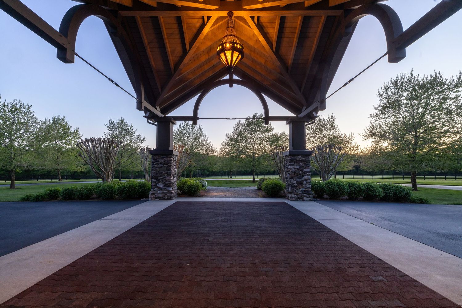 Elegant brick pavilion with wooden vaulted ceiling and stone columns at dusk, surrounded by lush green trees.