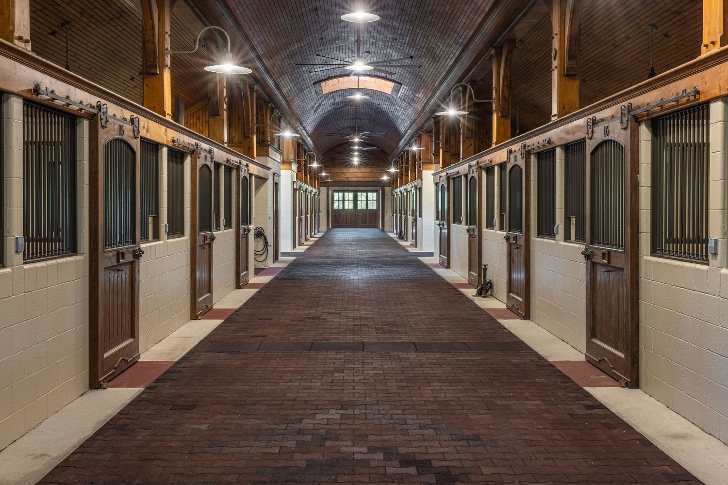 Elegant horse stable hallway with brick flooring, wooden stalls, and arched ceiling lighting in a well-maintained equestrian facility.