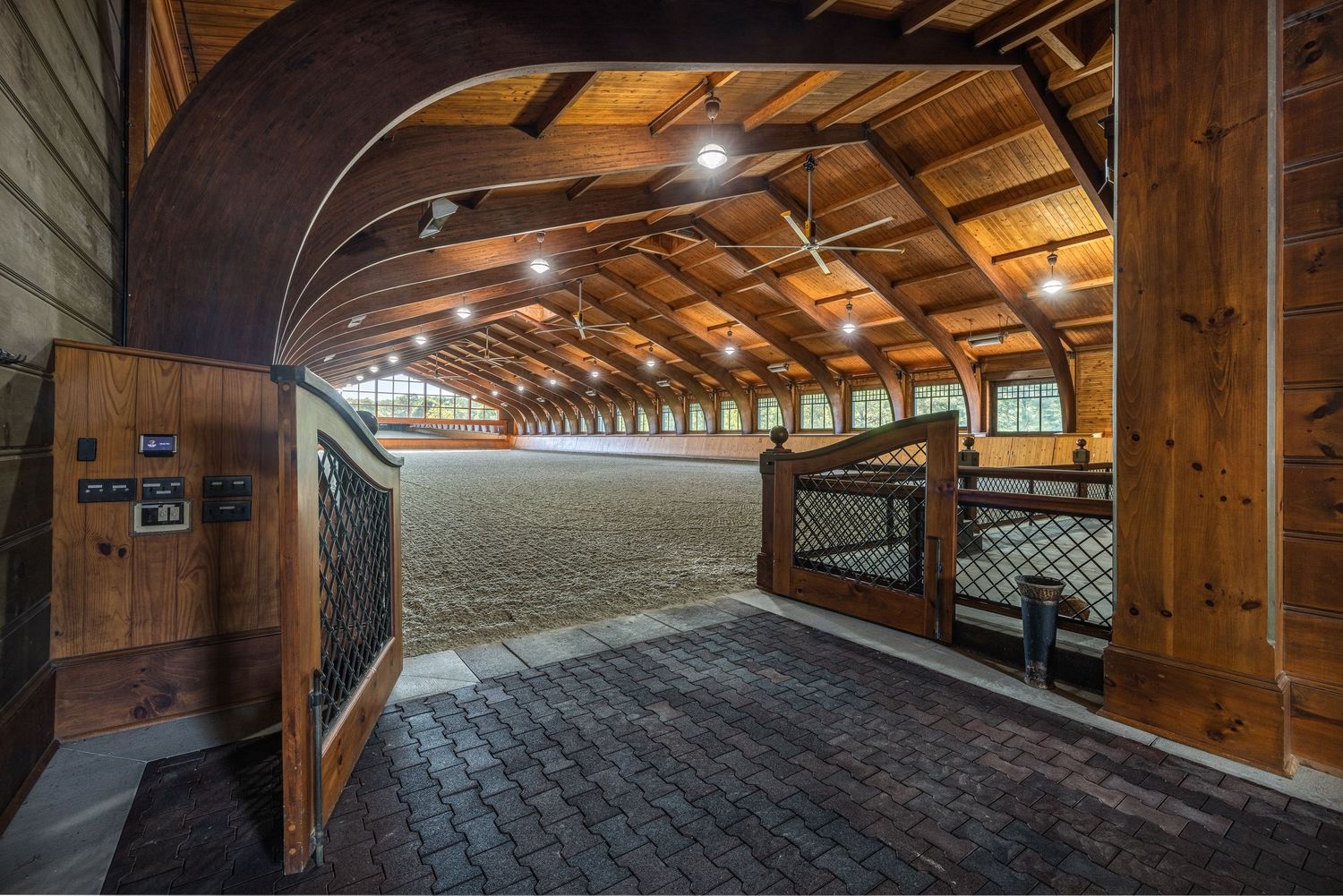 Elegant indoor equestrian arena with arched wooden ceiling, arena lighting, brick entry, and horse stalls in a luxury stable.
