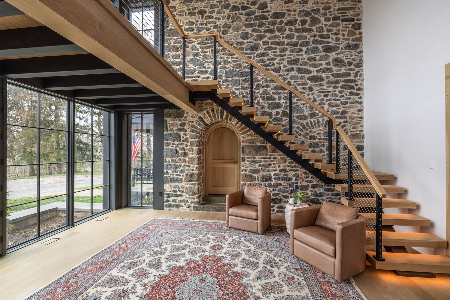 Modern home interior featuring a stone accent wall, floating staircase, leather chairs, and a decorative arched niche with large windows.