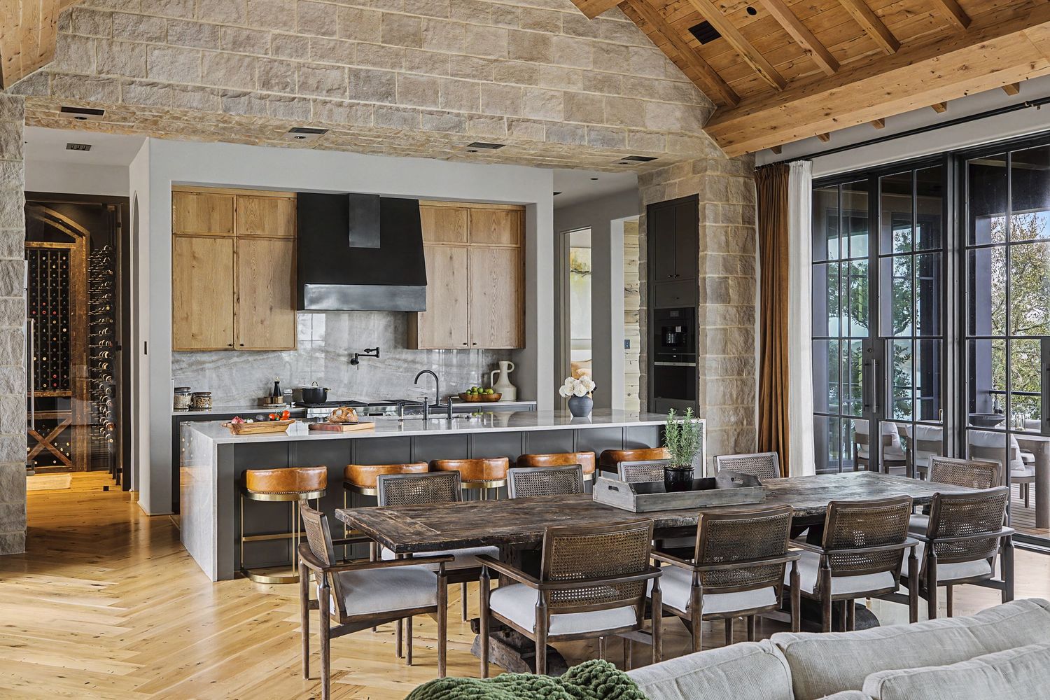 Rustic open-concept kitchen and dining area with stone walls, wood cabinets, herringbone floors, and large windows.