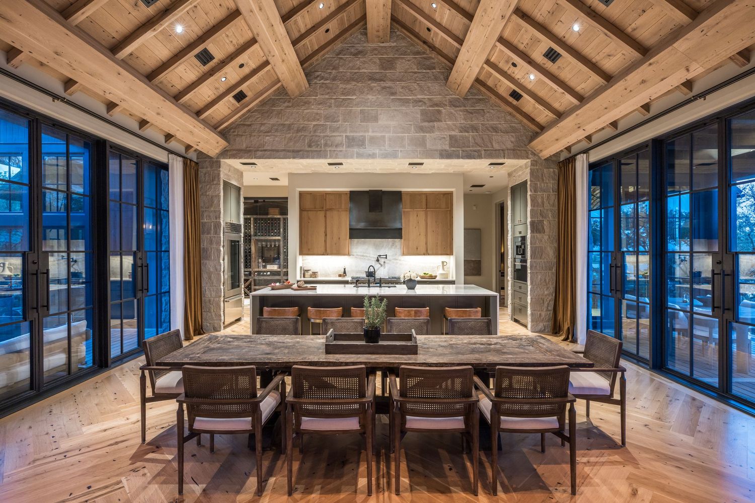 Luxury open-plan kitchen and dining area with vaulted wood beam ceiling, stone walls, and large glass doors.