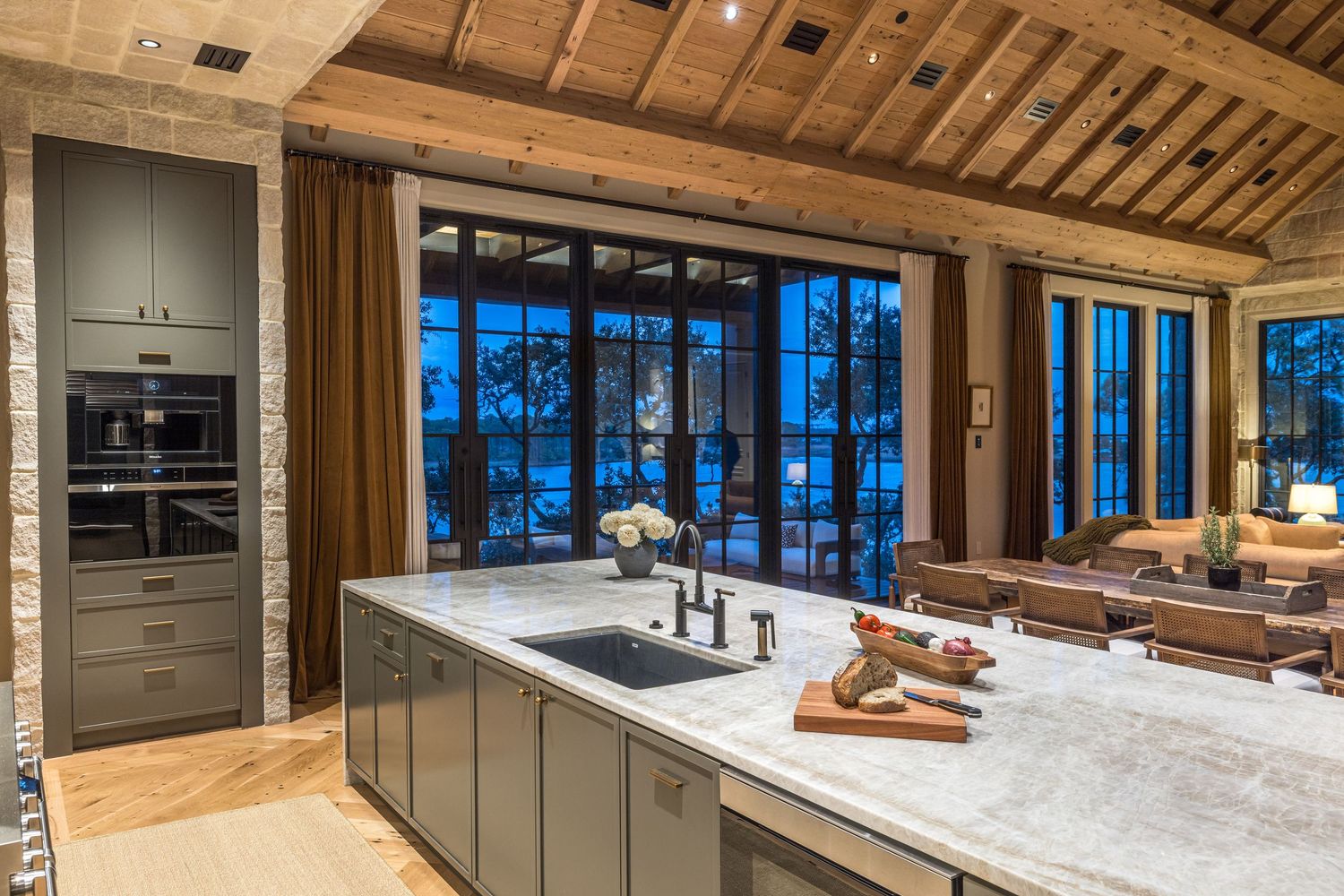 Luxury kitchen with marble island, gray cabinets, double ovens, and floor-to-ceiling windows with evening patio views.